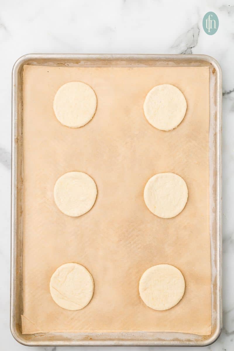 A baking sheet pan with parchment paper and 6 circle doughs.