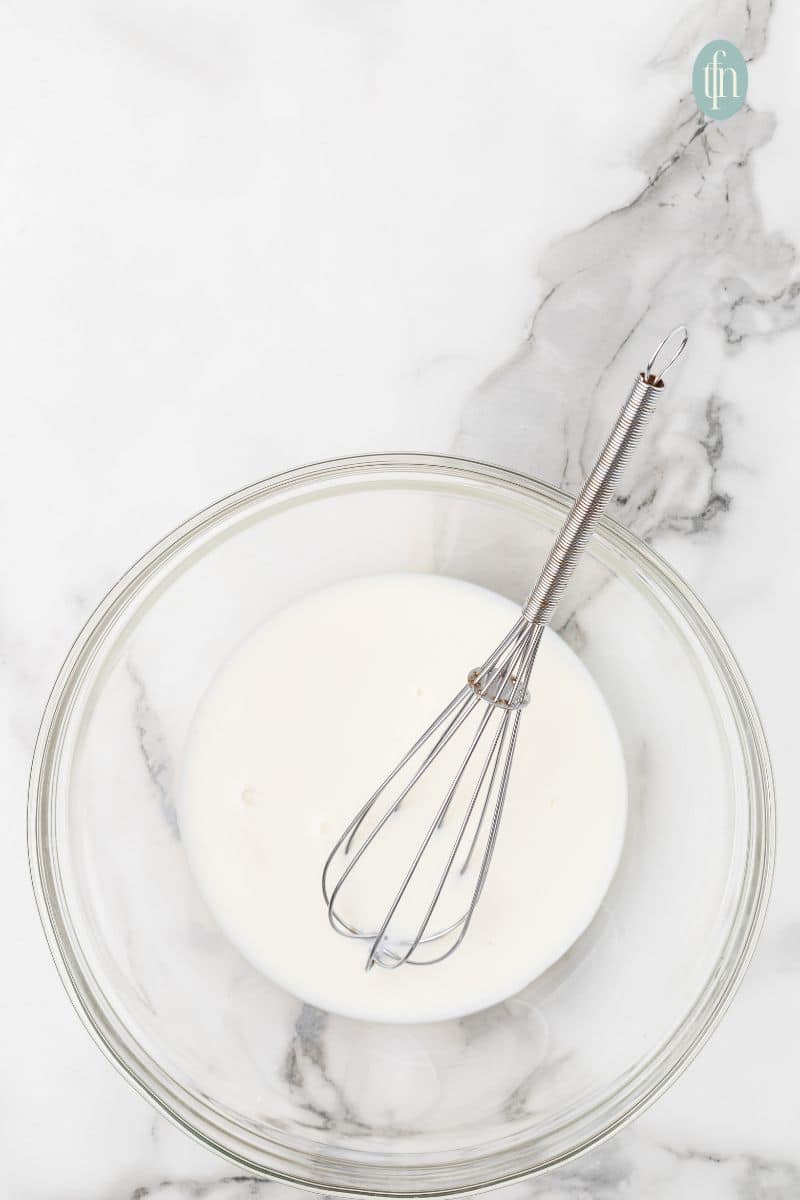 Mixed white liquid on a clear mixing bowl with whisk.