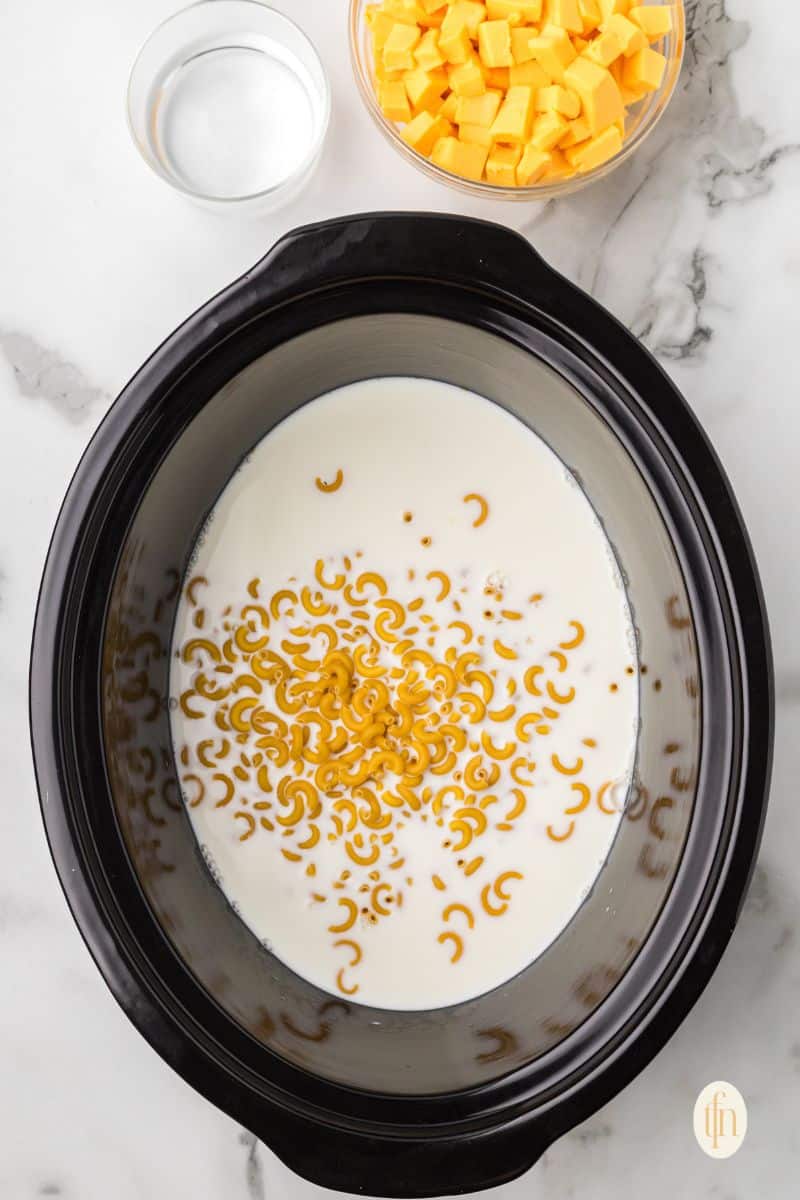 Crock pot with macaroni and milk and a bowl of water and cheese.