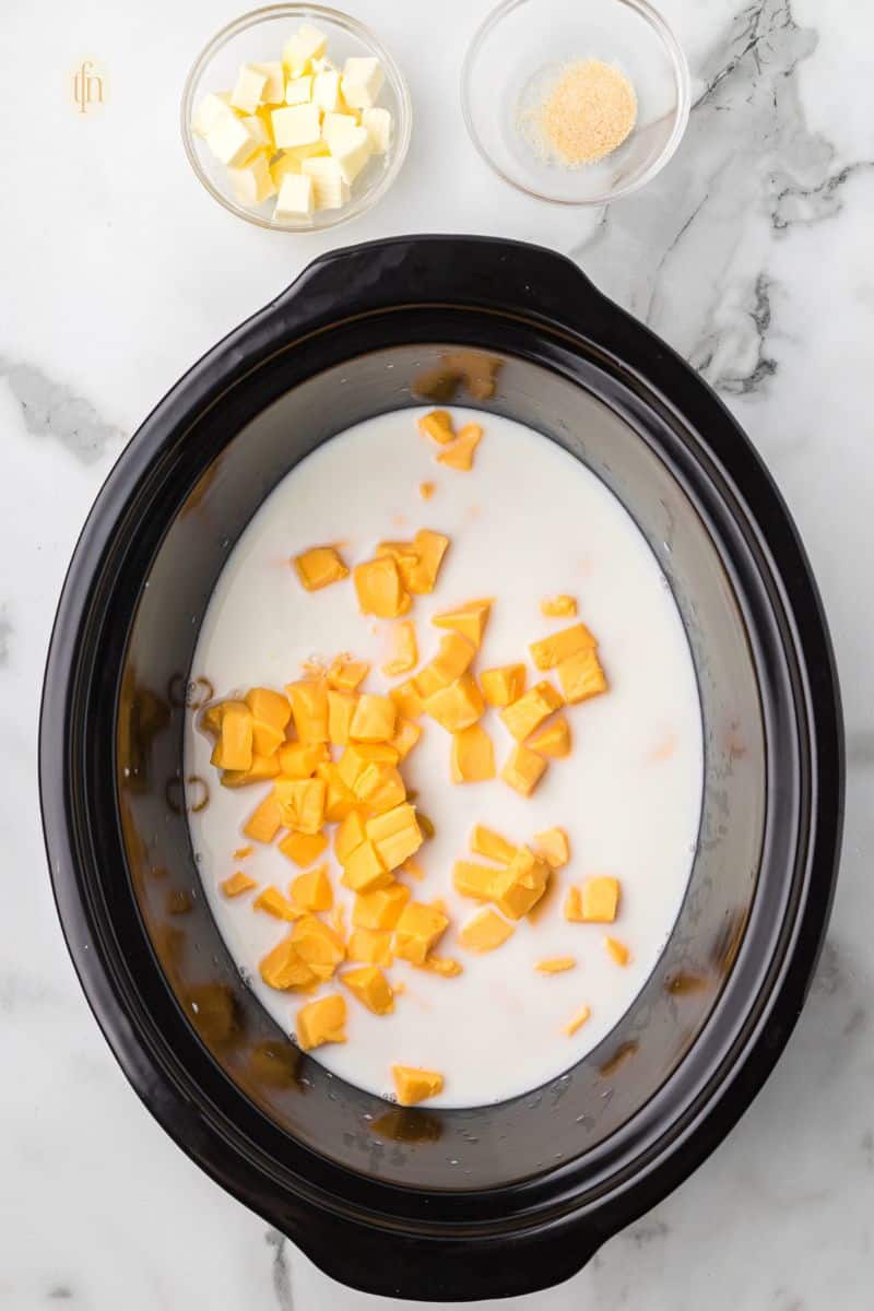 Crock pot with milk and cheese mixture and a bowl of butter and garlic powder.