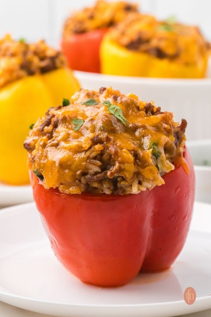An eye-level shot of a single baked red taco stuffed pepper on a white plate, with the top of a yellow pepper visible in the blurred background.