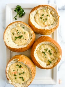 Four bread bowls filled with creamy soup garnished with fresh herbs, sitting on a white platter.
