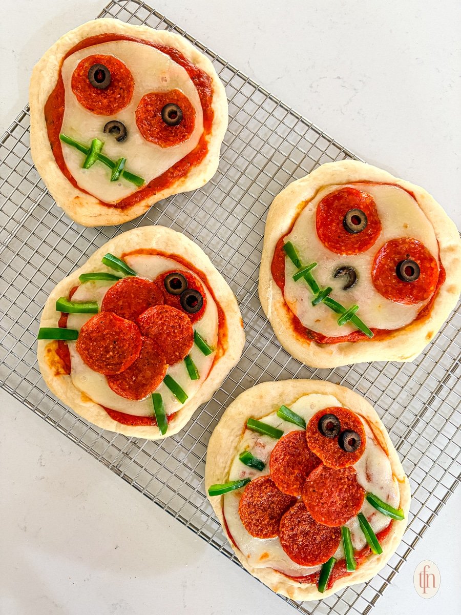 A group of four unbaked, Halloween-themed personal pizzas sits on a wire cooling rack. Two are decorated as skulls and two as spiders, using pepperoni, sliced olives, and green bell pepper strips.