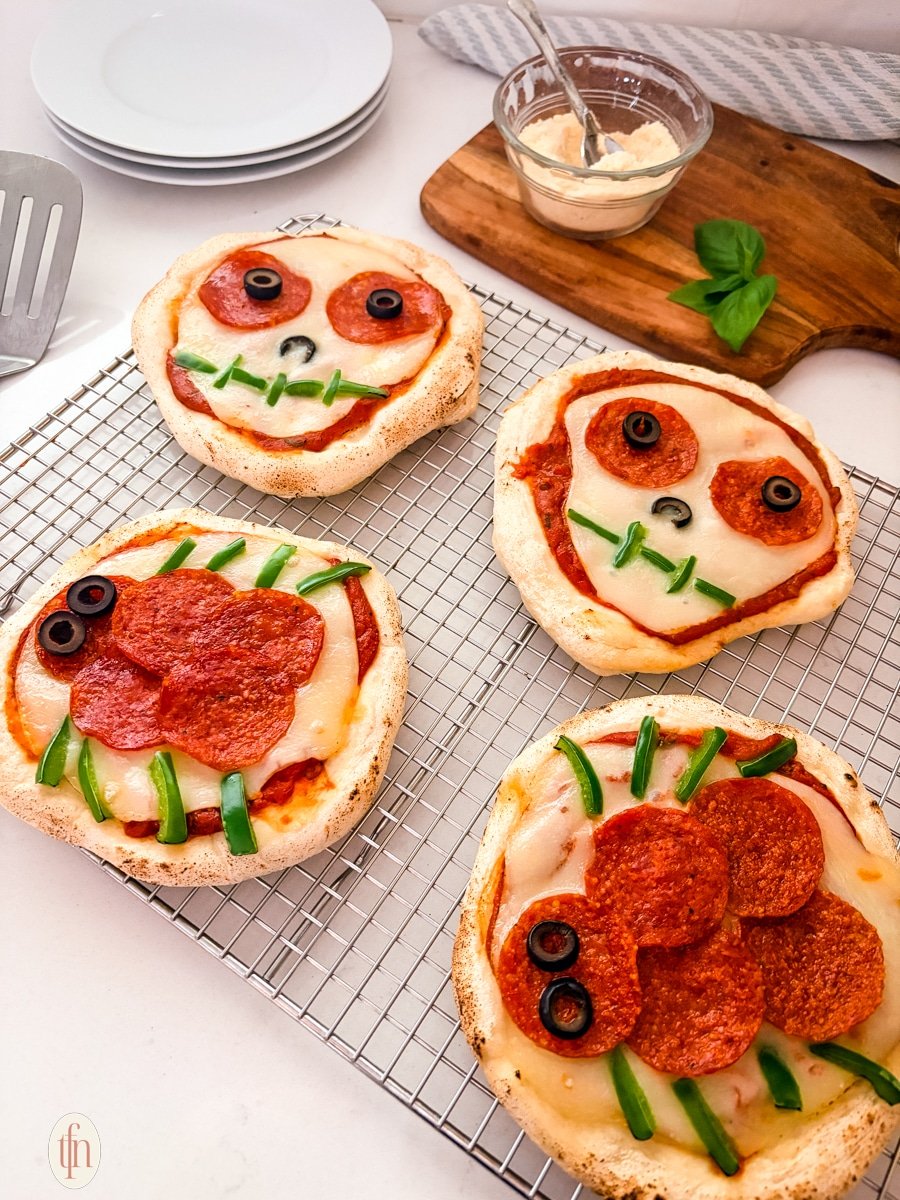 Four baked personal Halloween pizzas are shown on a wire cooling rack. Two are decorated as skulls with sliced olives for eyes and green peppers for a mouth, and two are decorated as spiders with pepperoni for the body and green pepper slices for legs.