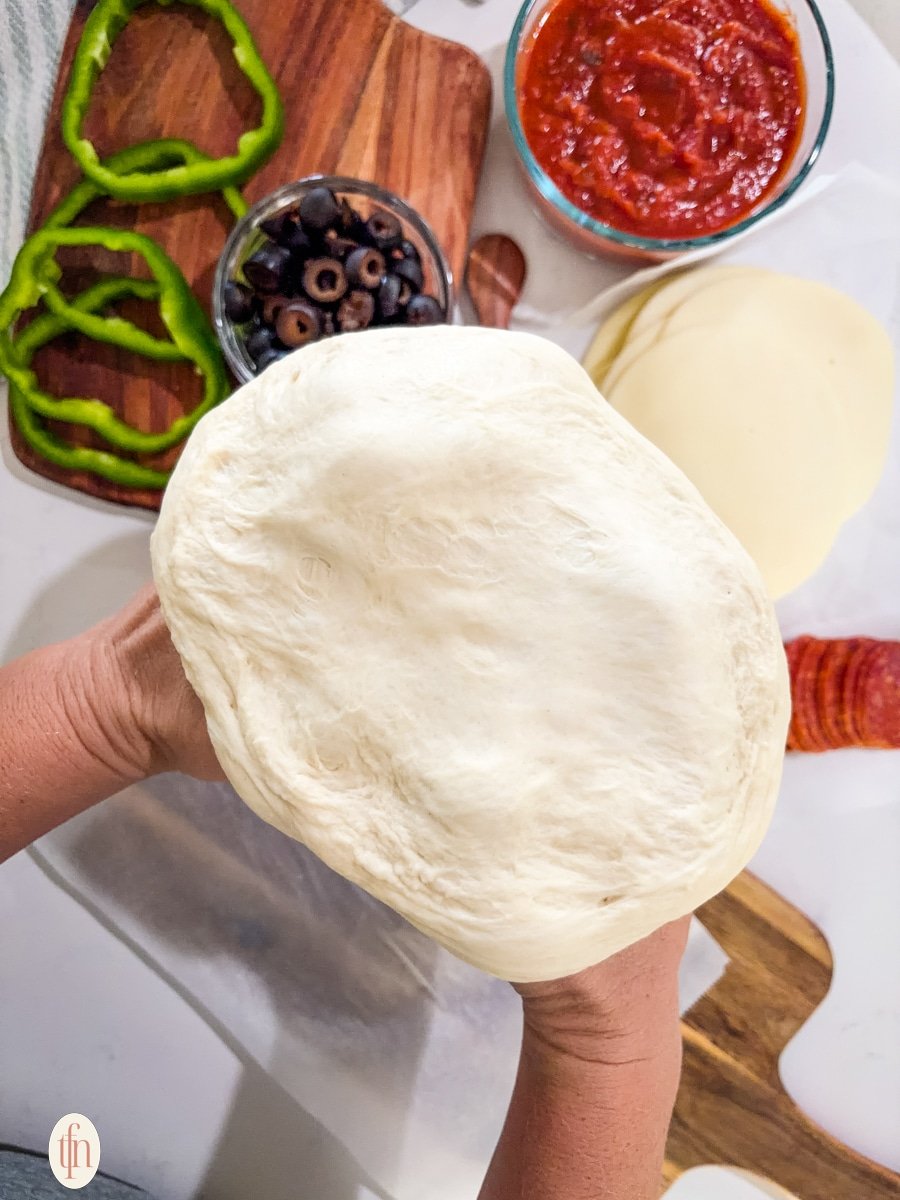 A pair of hands holds a portion of soft, unbaked pizza dough above a countertop with other pizza ingredients visible.