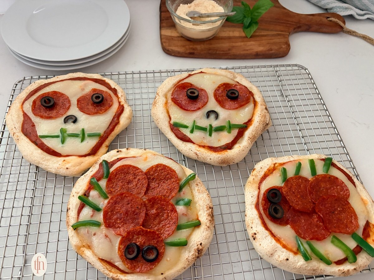 A wide, eye-level shot of four Halloween-themed personal pizzas on a wire cooling rack. Two are skulls and two are spiders, all made with pepperoni, olives, and green peppers.