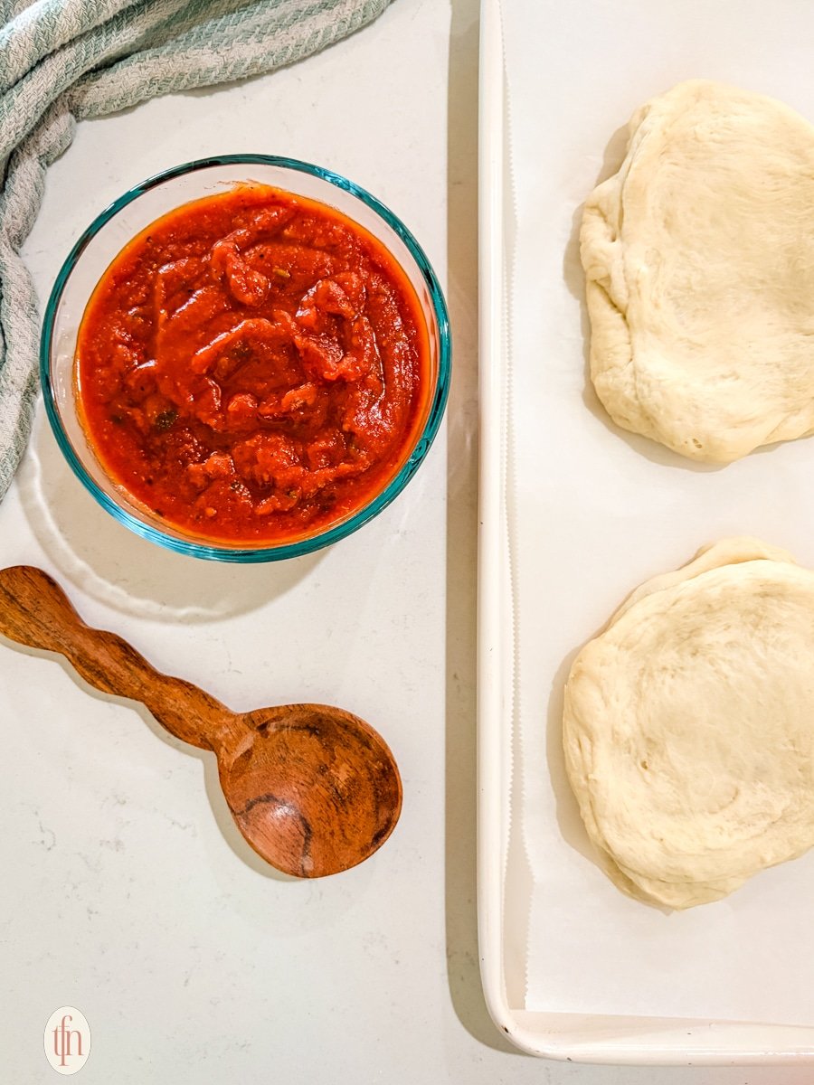 A bowl of marinara sauce and two rounds of pizza dough on a baking sheet lined with parchment paper are shown from above. A wooden spoon rests near the sauce bowl.