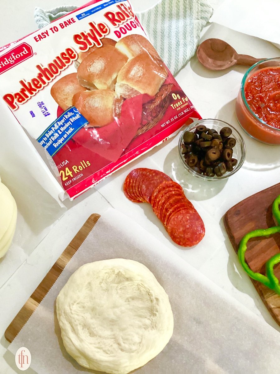 A bag of Parkerhouse Style Roll dough is prominently featured on a countertop surrounded by pizza ingredients like pepperoni, olives, and a small round of dough on parchment paper.