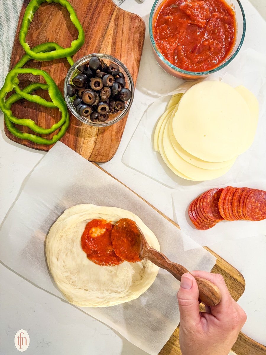 A hand uses a wooden spoon to spread red marinara sauce on a round of unbaked pizza dough. Sliced green peppers, olives, cheese, and pepperoni are arranged around it on the counter.