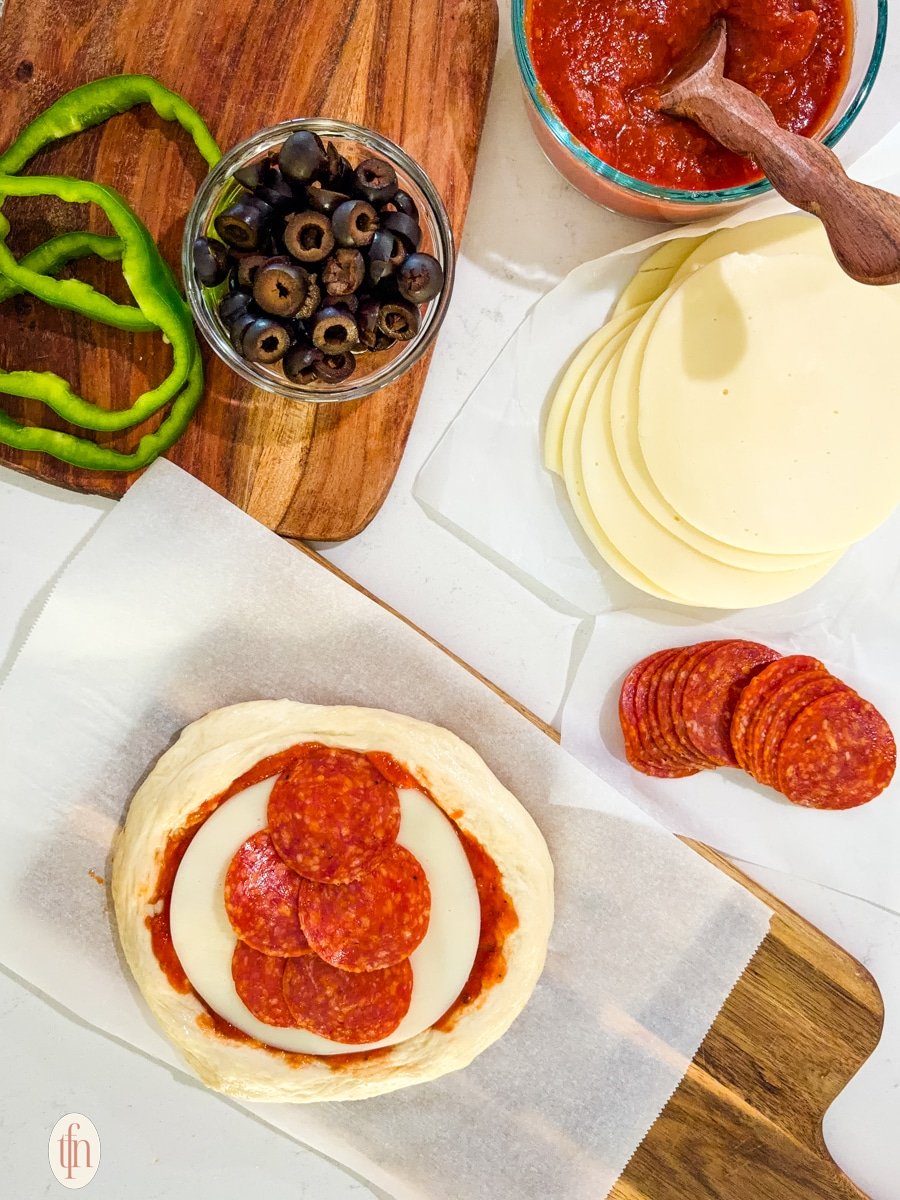 A round of unbaked pizza dough is topped with sauce, a slice of cheese, and several slices of pepperoni. Other ingredients are visible in the background.