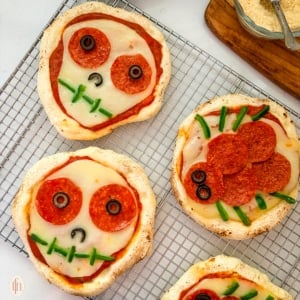 An overhead shot of four baked Halloween personal pizzas on a wire rack. Two are decorated as skulls with sliced olives for eyes and green peppers for a mouth, and two are decorated as spiders with pepperoni for the body and green pepper slices for legs.