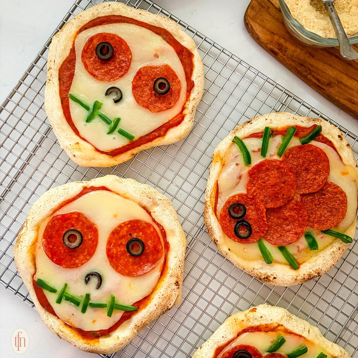 An overhead shot of four baked Halloween personal pizzas on a wire rack. Two are decorated as skulls with sliced olives for eyes and green peppers for a mouth, and two are decorated as spiders with pepperoni for the body and green pepper slices for legs.