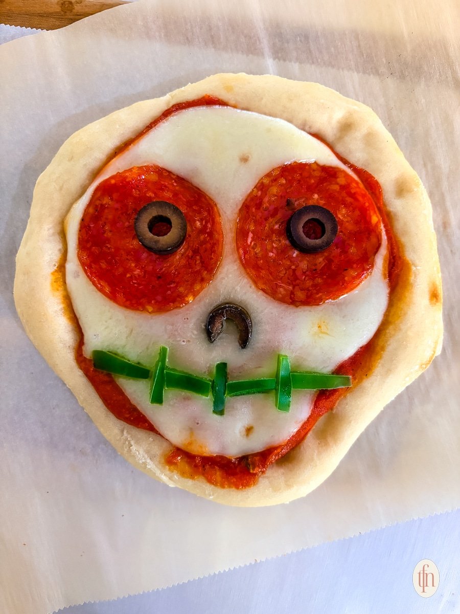 A close-up shot of an unbaked Halloween-themed personal pizza shaped like a skull. It has a marinara sauce base, mozzarella cheese, pepperoni for the eyes, sliced black olives for the pupils and nose, and small strips of green bell pepper for the mouth stitches.
