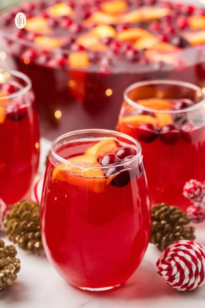 Close-up of glasses filled with vibrant red Christmas punch, garnished with orange slices and cranberries. A large punch bowl is blurred in the background, set with festive gold and red ornaments.