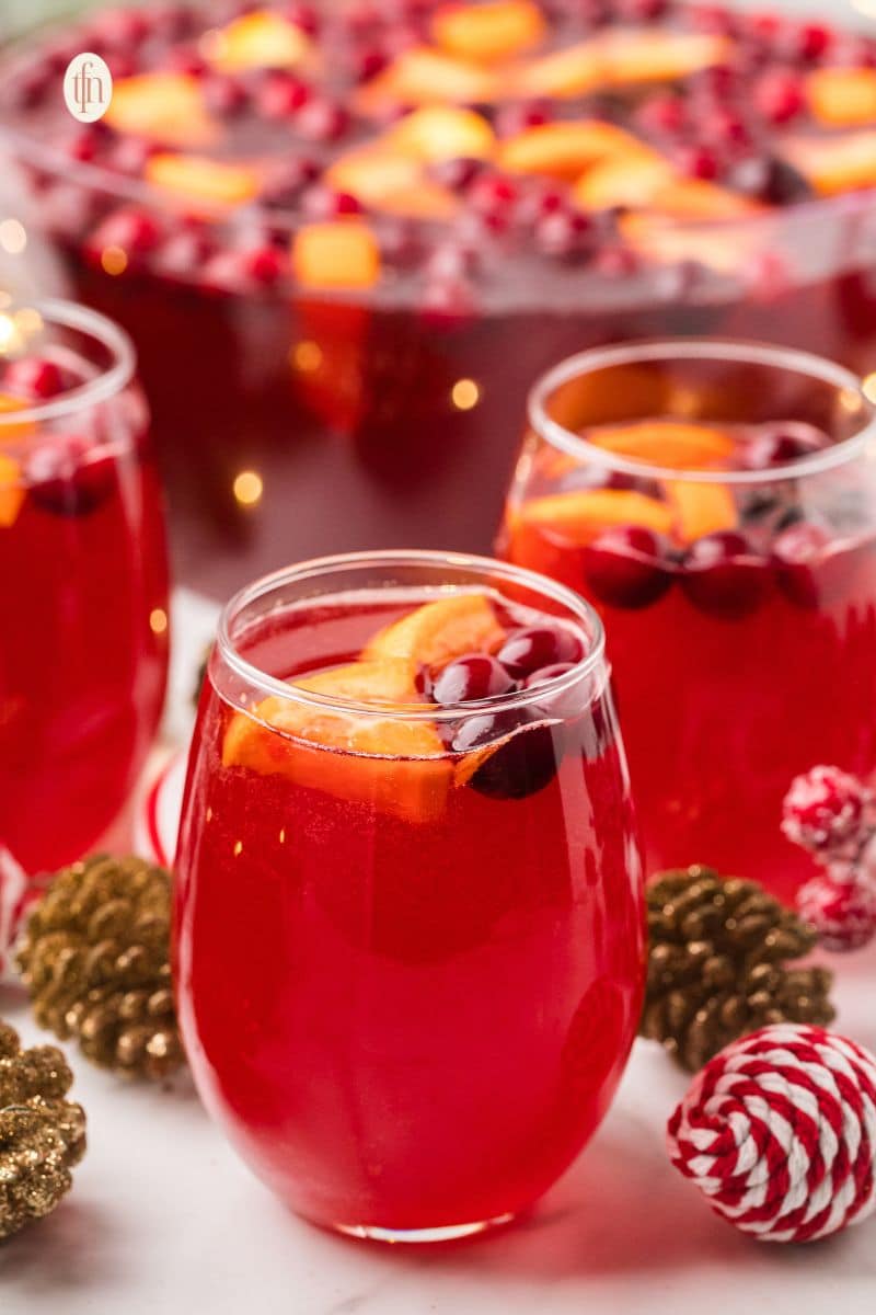 Close-up of glasses filled with vibrant red Christmas punch, garnished with orange slices and cranberries. A large punch bowl is blurred in the background, set with festive gold and red ornaments.