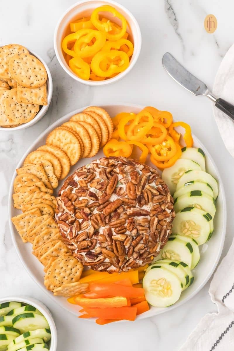 Overhead view of a completed pecan cheese ball appetizer on a platter, surrounded by circular rows of round crackers, sliced cucumbers, and yellow and orange bell pepper rings.