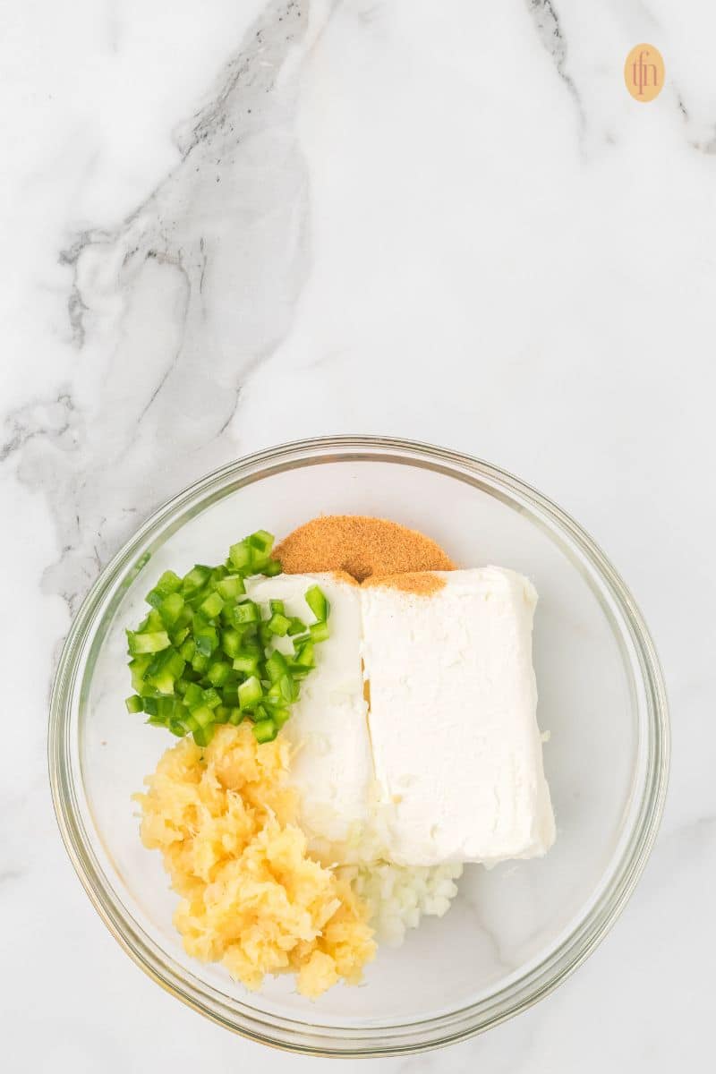 A clear glass bowl containing the raw ingredients for the cheese ball: a block of cream cheese, chopped green peppers, crushed pineapple, and spices/seasonings.