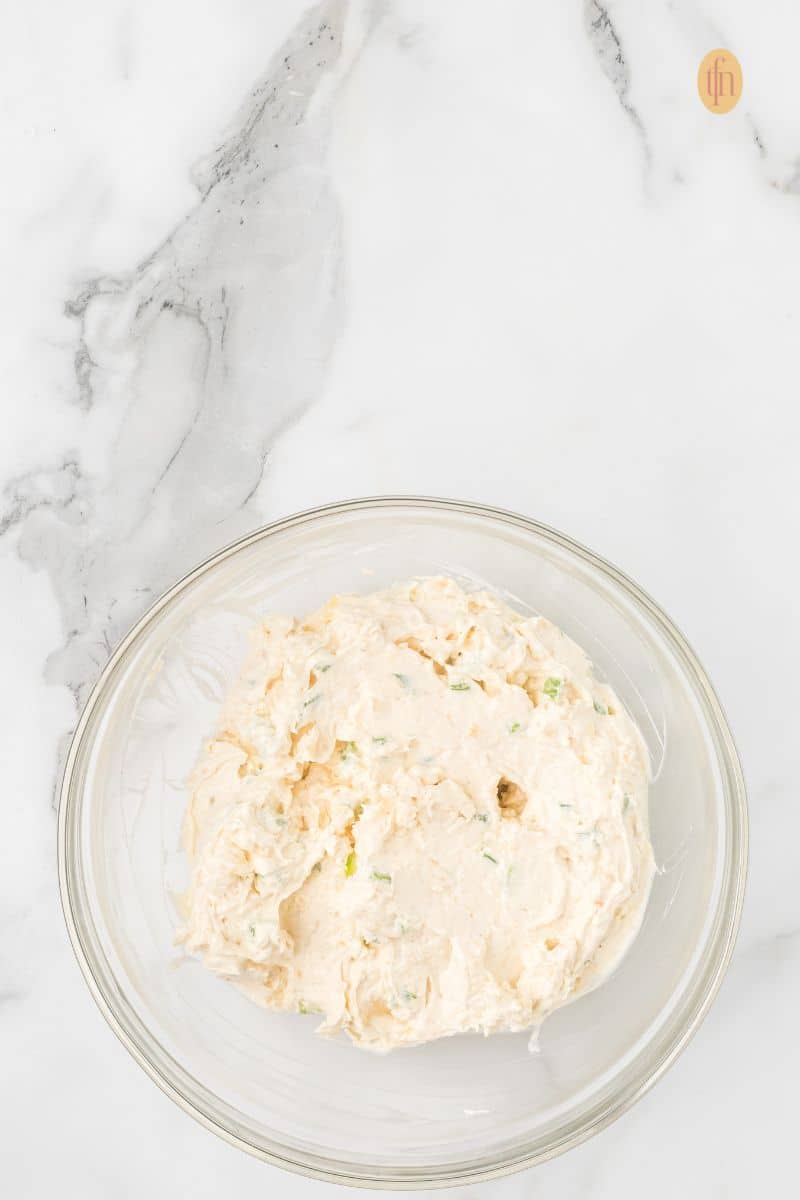 A clear glass bowl holding the fully mixed cheese ball ingredients—a creamy, off-white mixture with visible green pieces of seasoning or pepper.