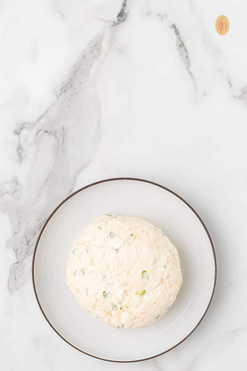 Top-down view of a round, smooth cream cheese ball (without a pecan coating) resting on a small white plate with a brown rim, set against a marble background.