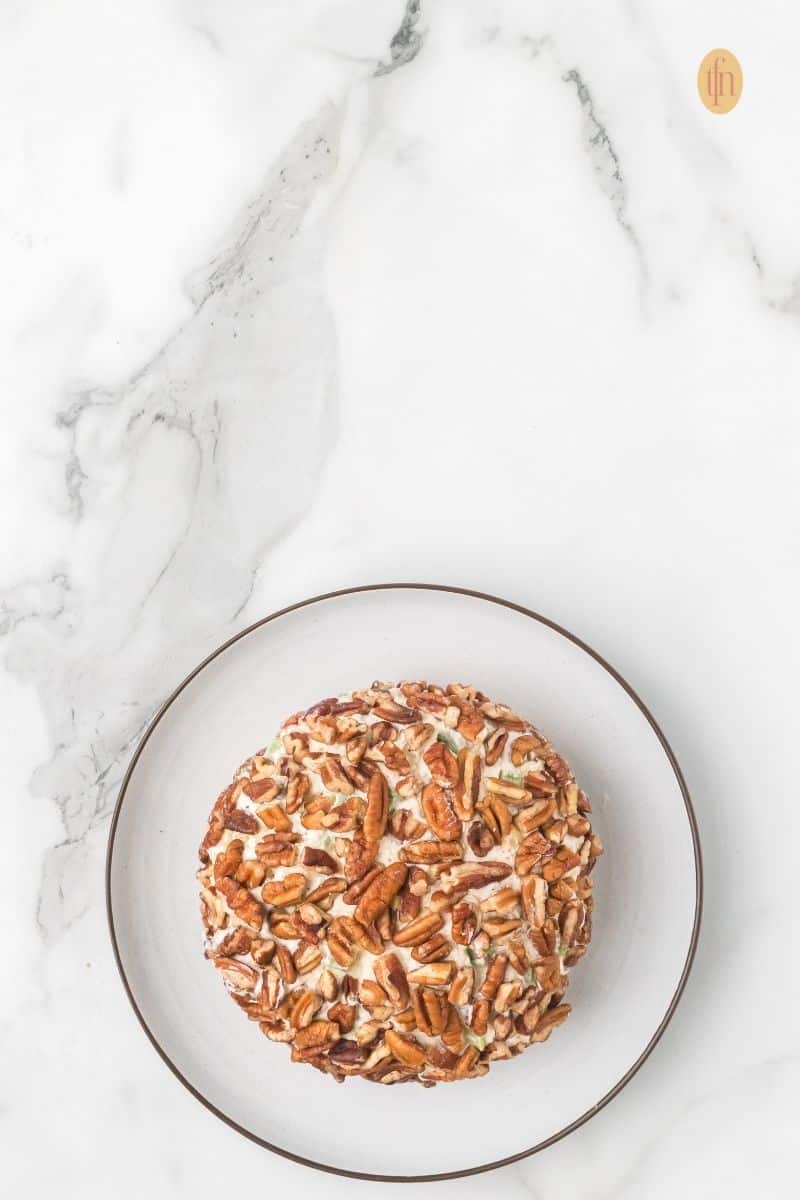 Top-down view of a cheese ball that has just been coated entirely with pecan halves, resting on a small white plate against a marble countertop.
