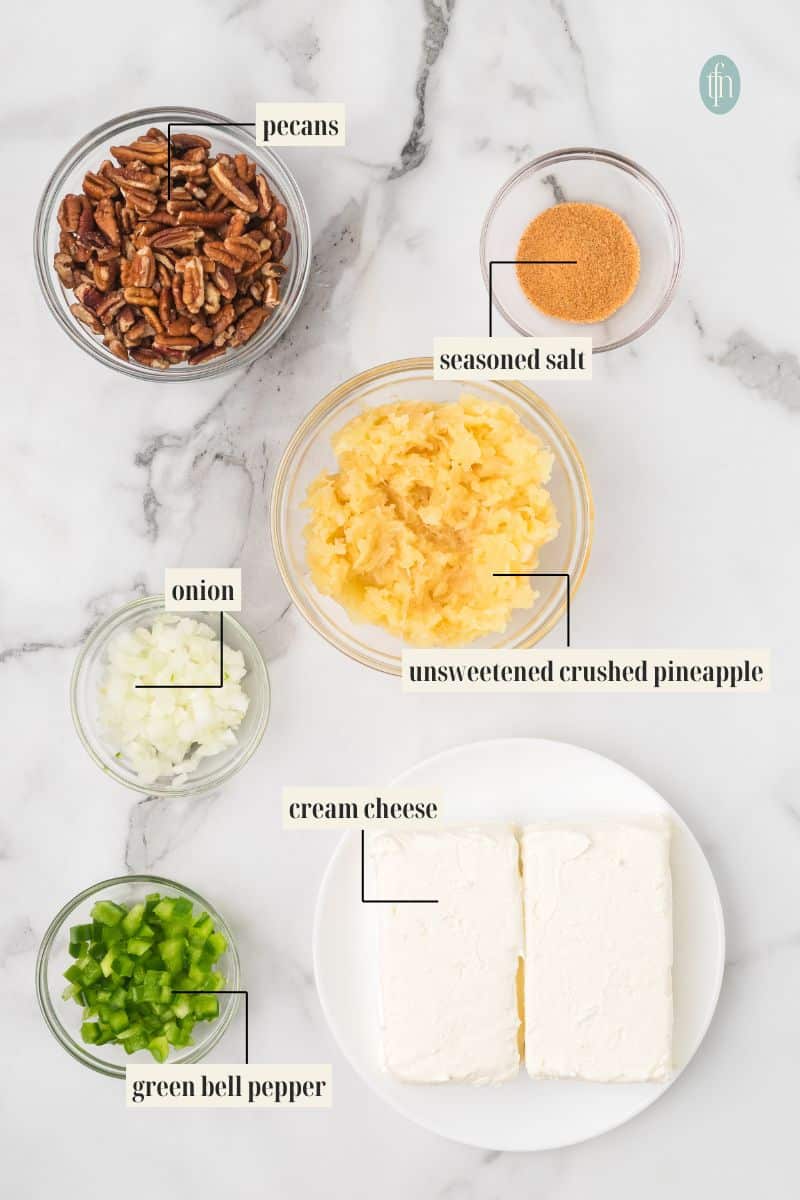 Overhead shot of all the ingredients needed to make a pineapple cheese ball laid out on a marble surface. Ingredients include pecans, seasoned salt, crushed pineapple, chopped onion, chopped green bell pepper, and two blocks of cream cheese.