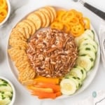 A round cream cheese ball coated entirely in pecans, served on a white platter with crackers, sliced cucumbers, and rings of yellow and orange bell peppers.