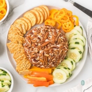 A round cream cheese ball coated entirely in pecans, served on a white platter with crackers, sliced cucumbers, and rings of yellow and orange bell peppers.