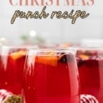 Close-up of glasses filled with vibrant red Christmas punch, garnished with orange slices and cranberries. A large punch bowl is blurred in the background, set with festive gold and red ornaments.