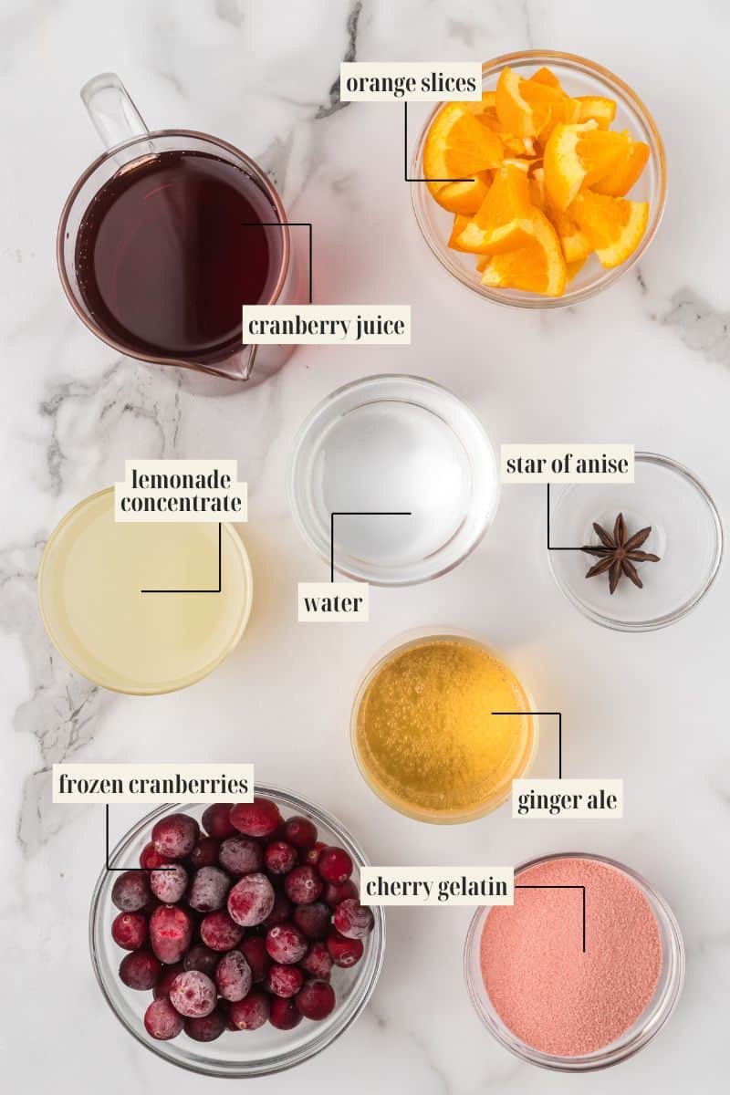 Flat lay photo of ingredients for holiday punch: a pitcher of cranberry juice, bowls of orange slices, frozen cranberries, lemonade concentrate, water, ginger ale, cherry gelatin mix, and a star of anise.
