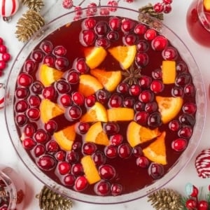 Overhead view of a large glass bowl of holiday punch garnished with floating cranberries, orange slices, and a star of anise. The bowl is surrounded by Christmas decor like gold pinecones and red bead garlands.