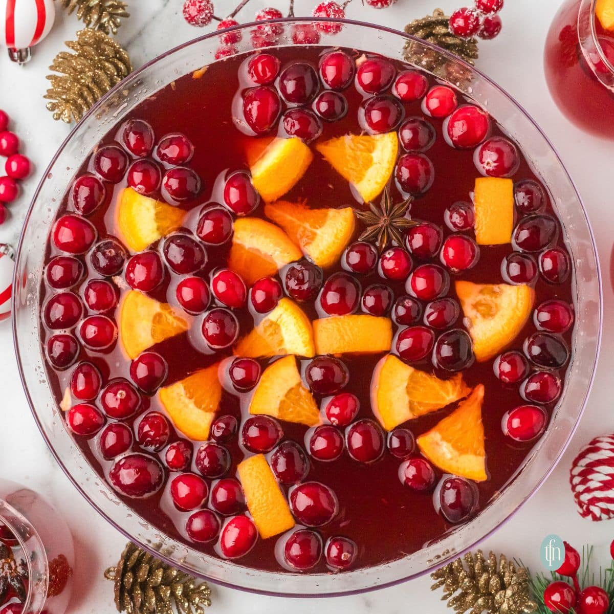 Overhead view of a large glass bowl of holiday punch garnished with floating cranberries, orange slices, and a star of anise. The bowl is surrounded by Christmas decor like gold pinecones and red bead garlands.