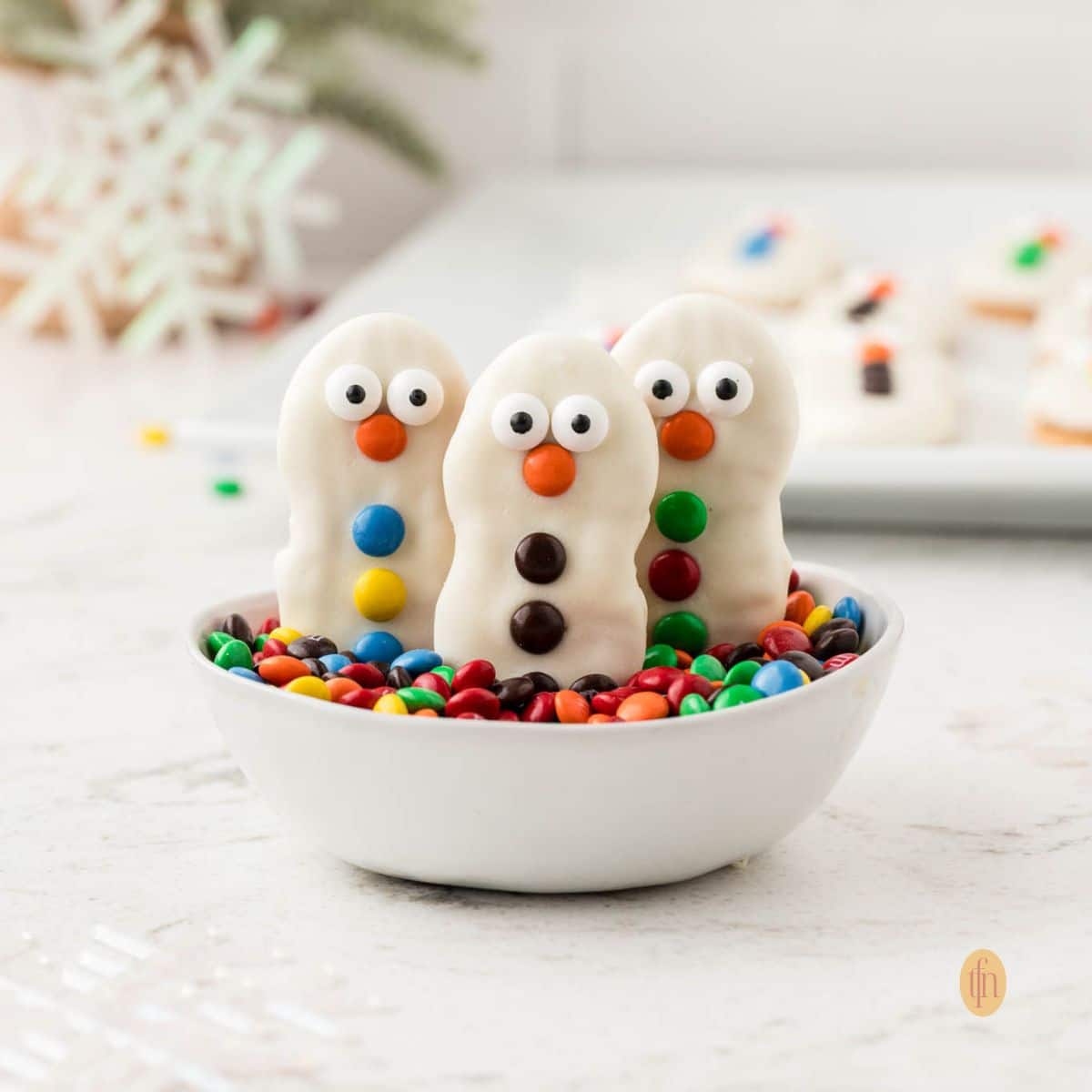 A close-up, square-cropped shot of three decorated Nutter Butter snowman cookies standing upright in a small white bowl filled with colorful M&M candies. A snowflake decoration is blurred in the background.