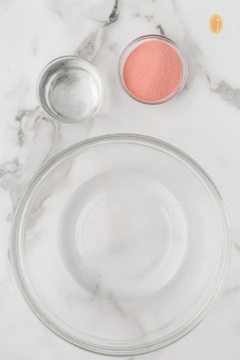 Overhead view of the initial punch ingredients: an empty mixing bowl, a small bowl of water, and a small bowl of cherry gelatin powder on a white marble surface.