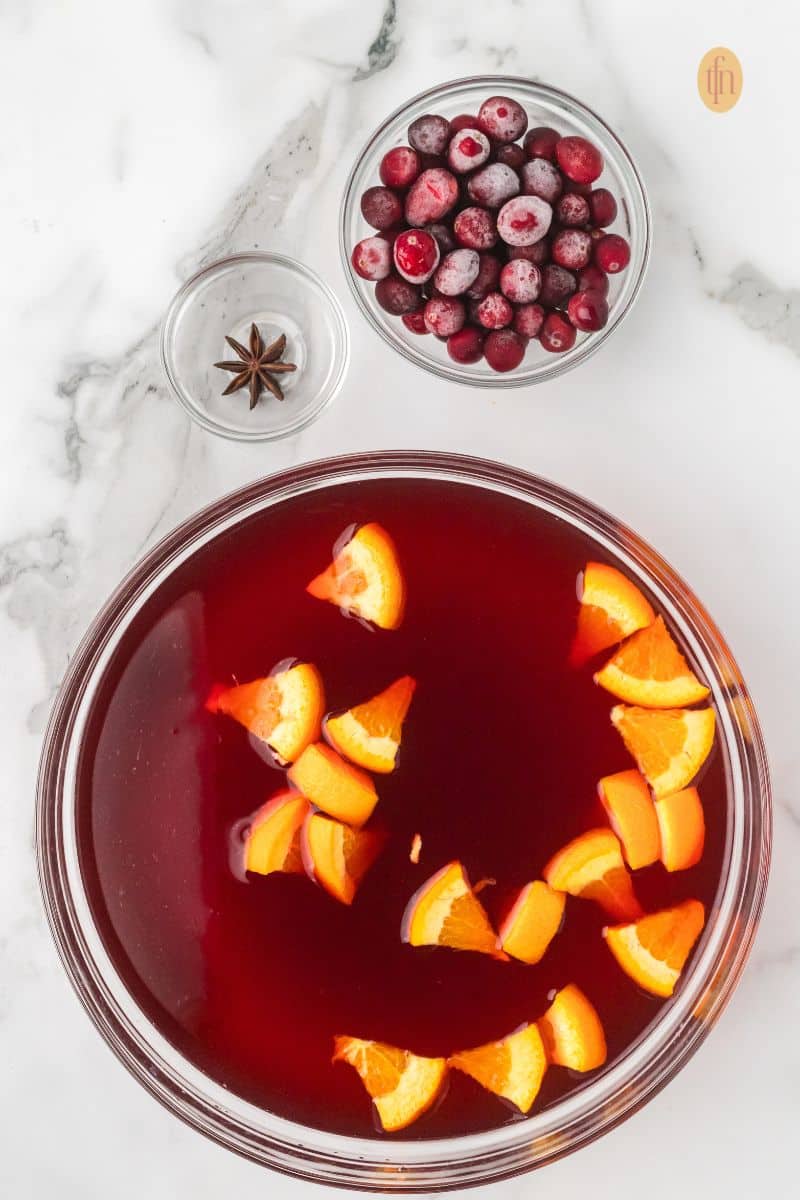 Ingredients for punch: a glass bowl of red liquid with orange slices, a bowl of frozen cranberries, and a small bowl with a star of anise on a marble counter.