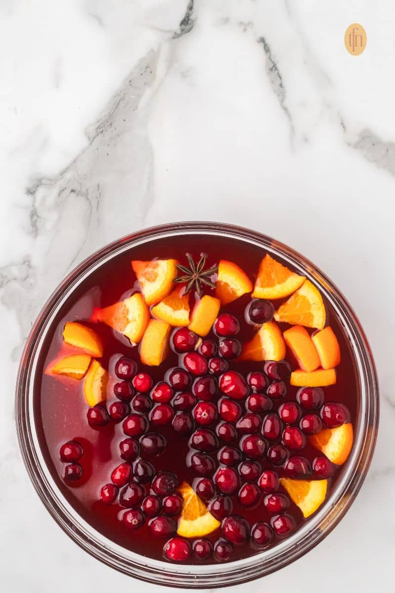 Overhead view of a punch bowl filled with red punch, currently being garnished with cranberries, orange slices, and star anise before serving.