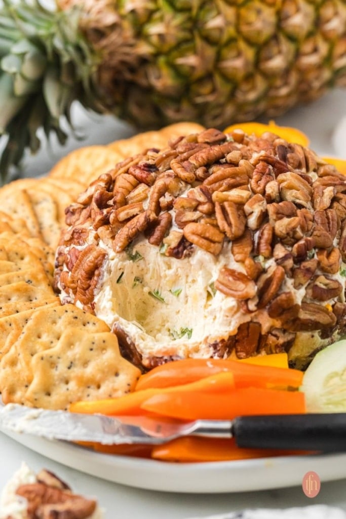 Close-up of a serving platter with a pecan-crusted cheese ball that has a scoop taken out, revealing the creamy pineapple and herb filling. A cracker and a knife are visible in the foreground.