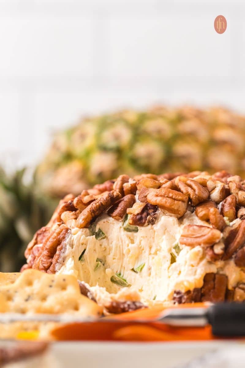 A side view close-up of the pecan cheese ball where a piece has been scooped out, showing the light-colored, herbed filling. A butter knife and a cracker are visible in the foreground, and a pineapple is blurred behind it.