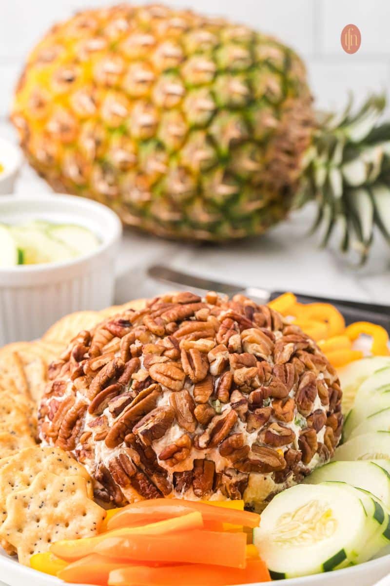 The pecan-crusted cheese ball appetizer served on a platter with sliced cucumbers and orange bell peppers in the foreground, with a whole fresh pineapple sitting on the counter in the background.