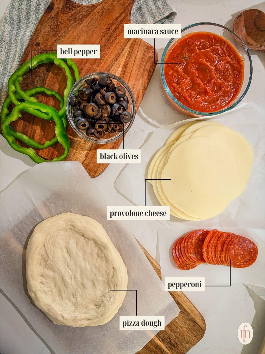 An overhead shot of the ingredients for Halloween pizzas with each item labeled. The labels point to a round of pizza dough, a bowl of marinara sauce, sliced bell pepper, sliced black olives, rounds of provolone cheese, and a stack of pepperoni.