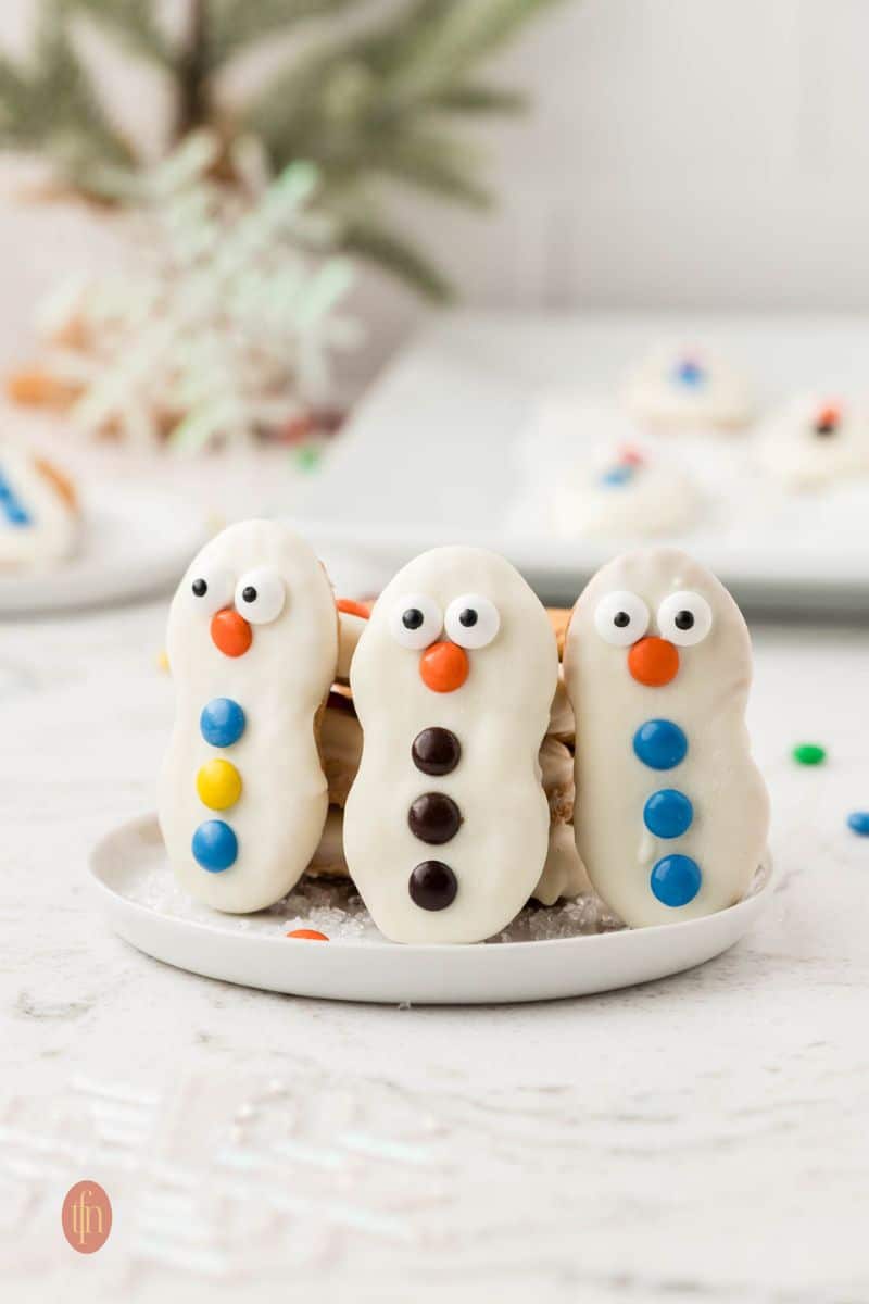 A close-up, eye-level shot of three decorated Nutter Butter snowman cookies standing upright on a small white plate dusted with sugar crystals.