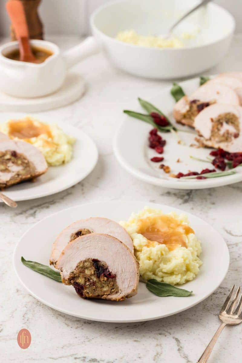A low-angle shot showing three plates of food on a marble counter. The foreground plate holds two slices of stuffed turkey breast and mashed potatoes with gravy. A gravy boat and a bowl of mashed potatoes are in the background.