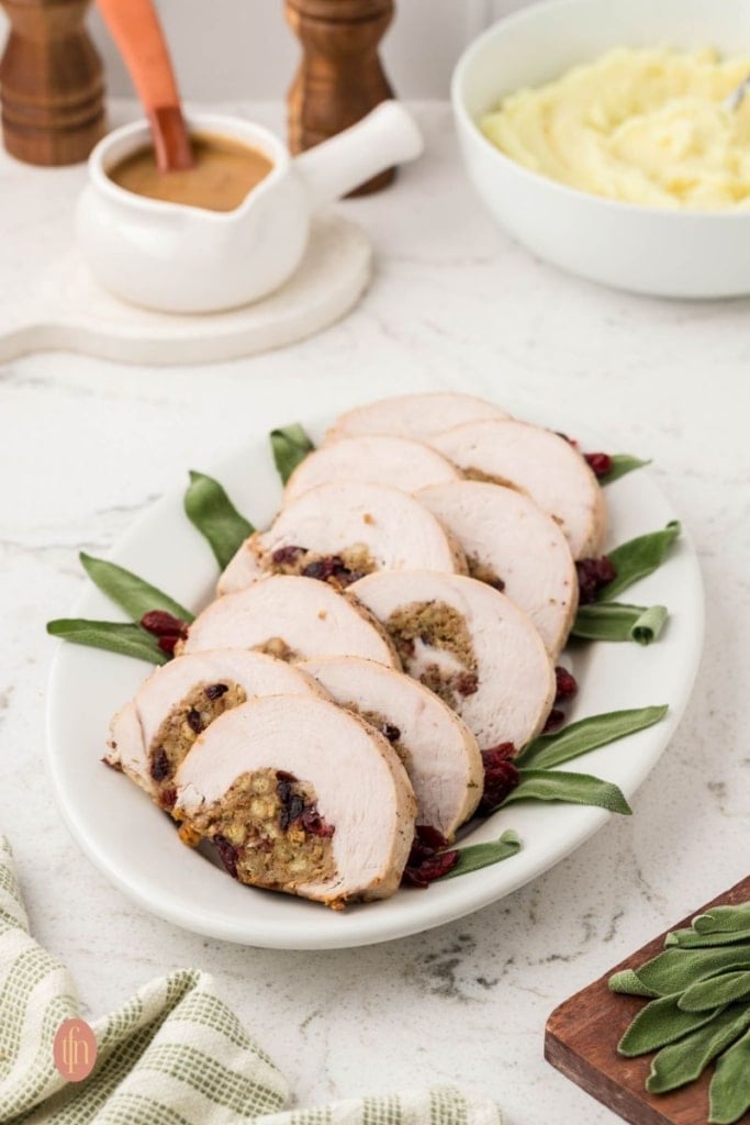 A wide, eye-level shot of a long white platter with seven slices of stuffed turkey breast, garnished with sage and dried cranberries. A gravy boat and a bowl of mashed potatoes are in the soft-focus background.