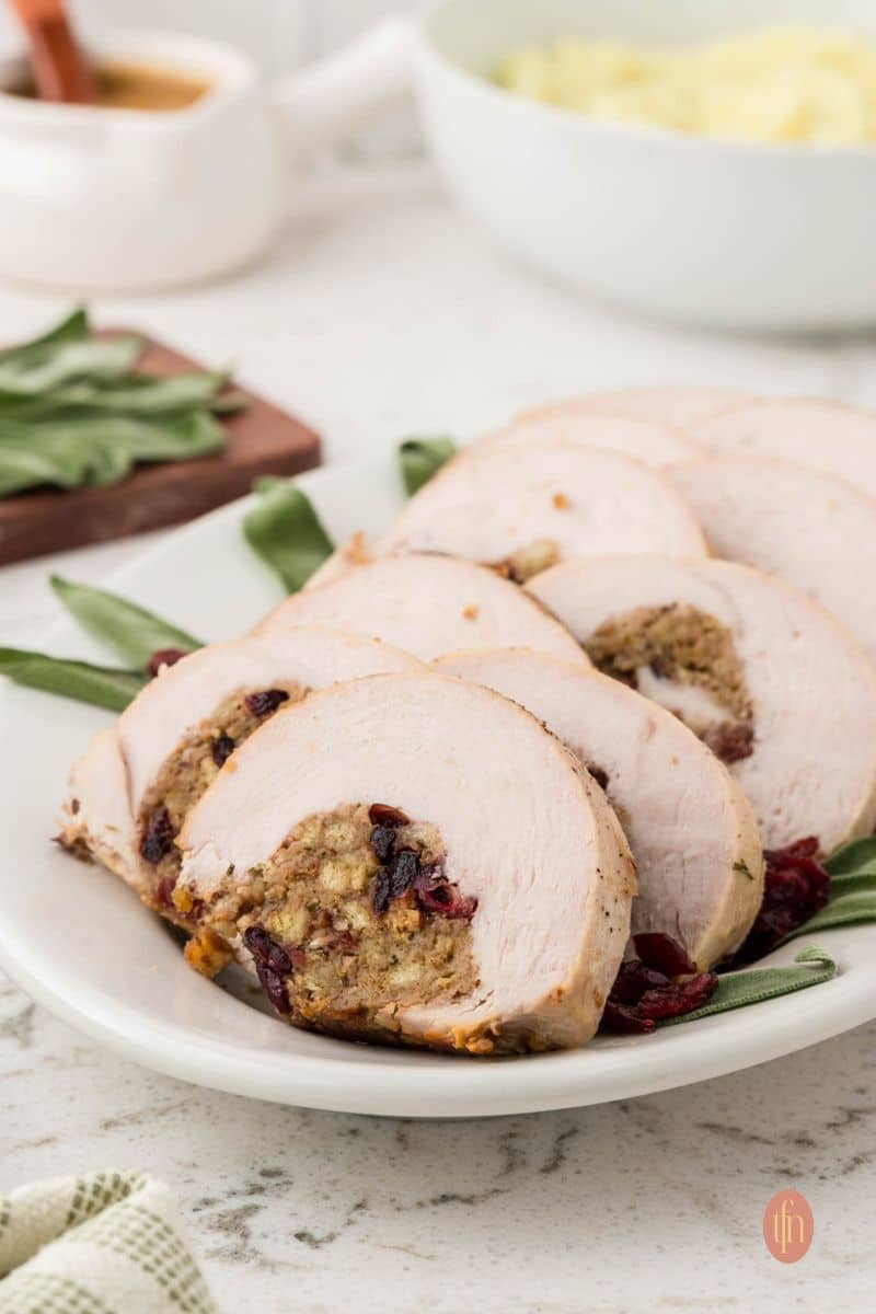 A slightly angled, eye-level shot of a long white platter with seven slices of stuffed turkey breast garnished with sage leaves and dried cranberries. A gravy boat and bowl of mashed potatoes are in the background.