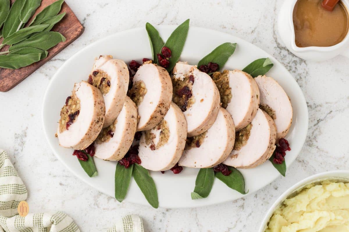 A wide, overhead shot of a long white platter holding slices of baked, stuffed turkey breast garnished with fresh sage leaves and dried cranberries. A gravy boat and a bowl of mashed potatoes are visible in the top and bottom corners.