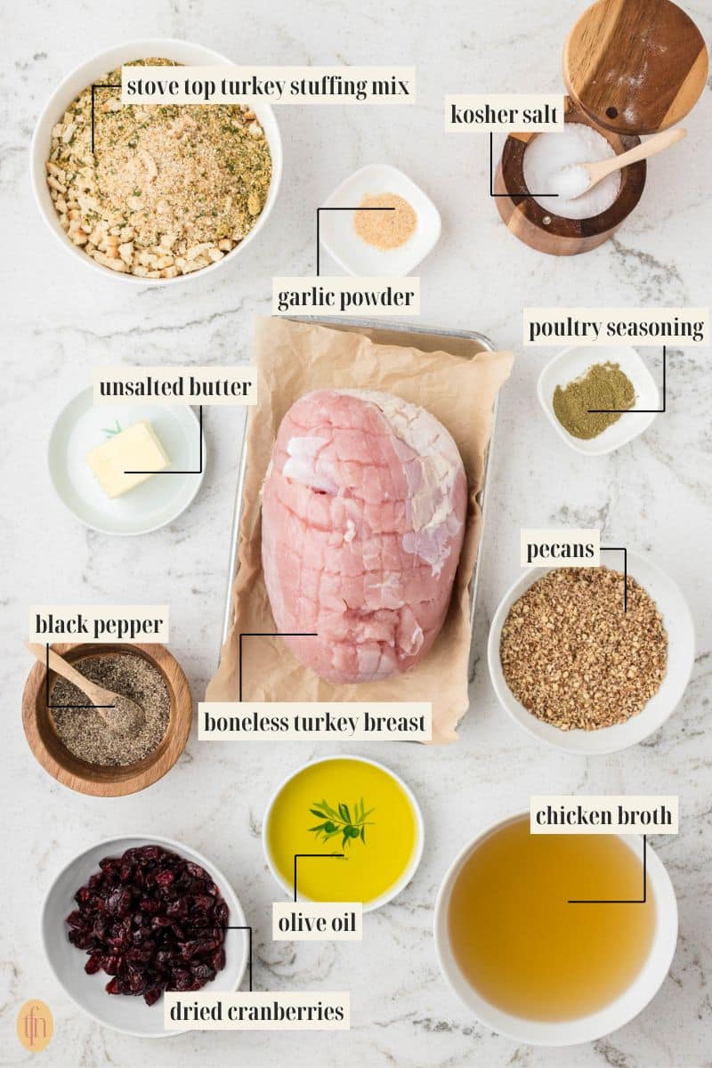 An overhead shot of all the ingredients for Stuffed Turkey Breast with each item clearly labeled: a bowl of stove top turkey stuffing mix, a slab of unsalted butter, black pepper, dried cranberries, garlic powder, a piece of raw boneless turkey breast, olive oil, ground pecans, poultry seasoning, kosher salt, and chicken broth.