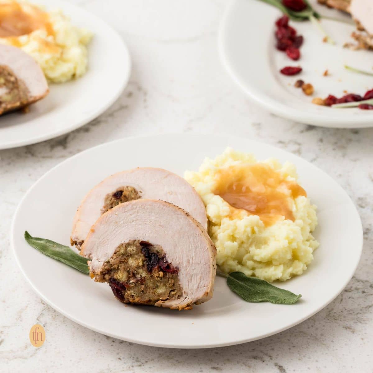 A square-cropped image of two slices of stuffed turkey breast served on a white plate next to a mound of mashed potatoes topped with gravy and garnished with a sage leaf.