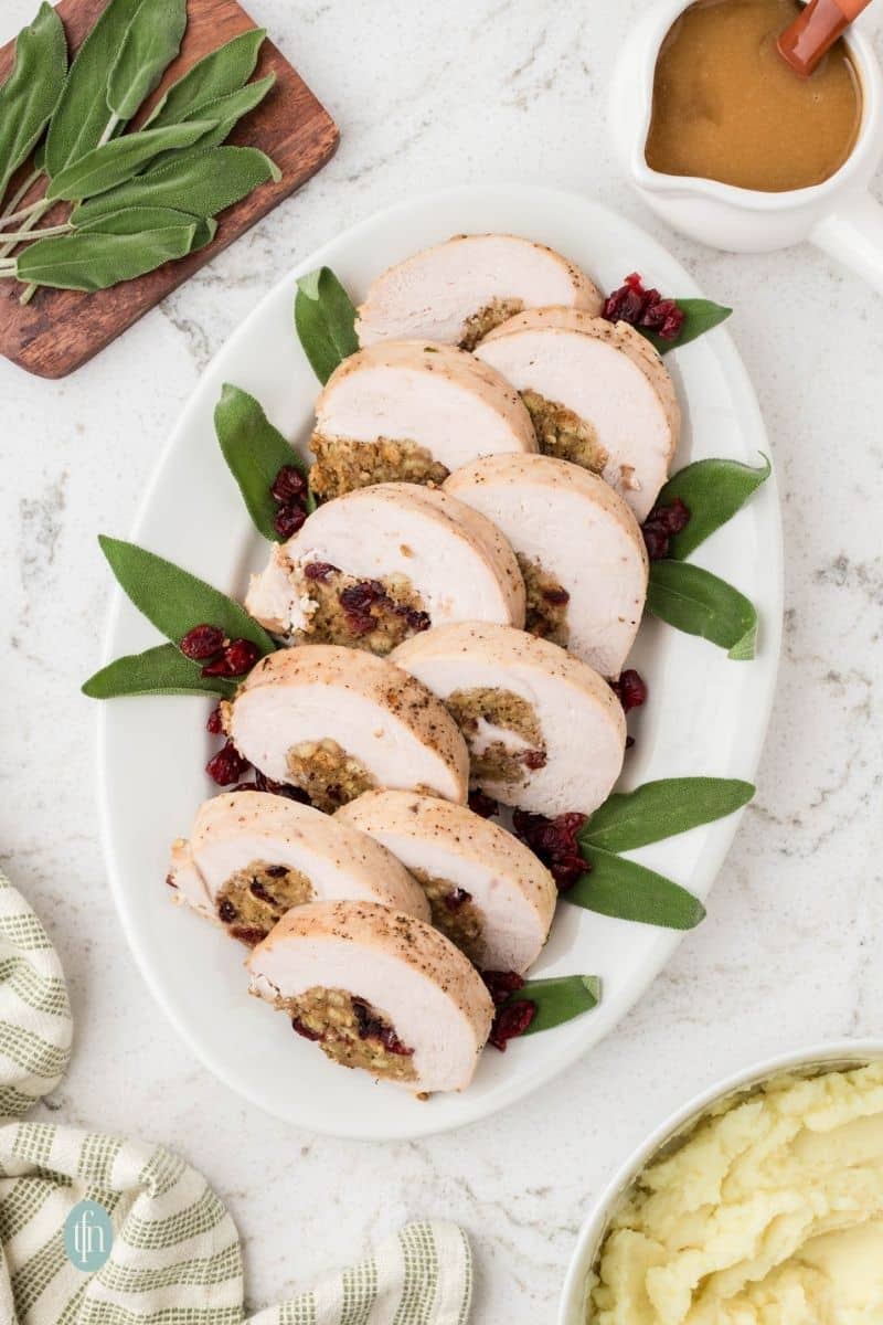 An overhead shot of a long white platter holding seven slices of baked, stuffed turkey breast garnished with fresh sage leaves and dried cranberries. A gravy boat and bowl of mashed potatoes are visible on the counter.