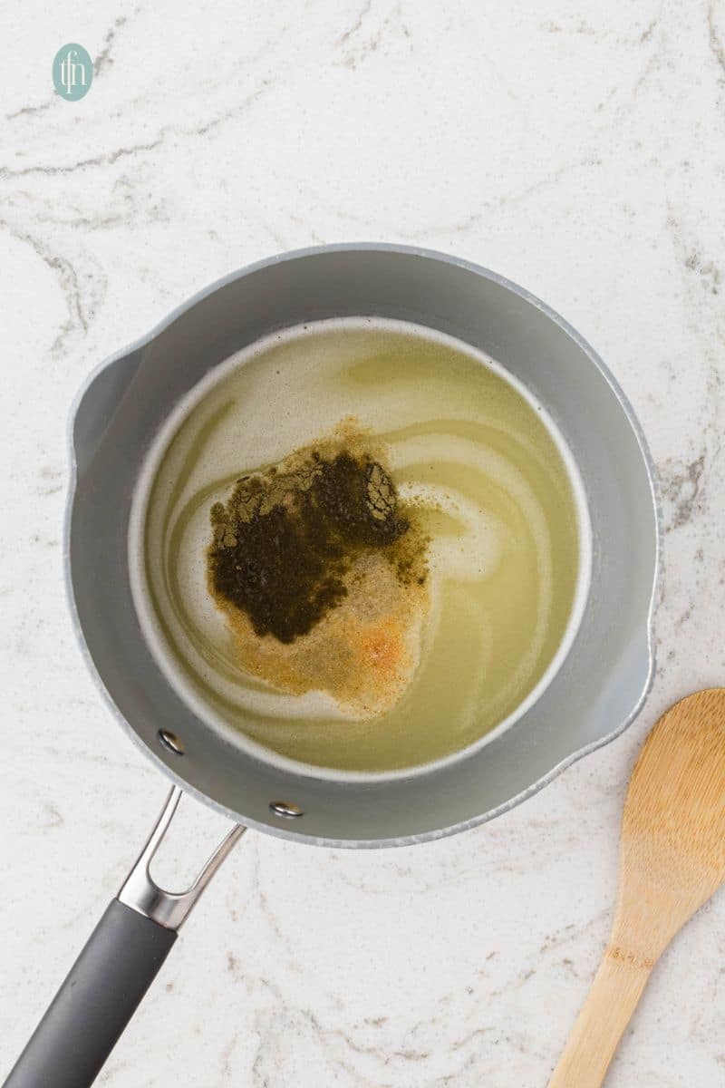 An overhead shot of a saucepan containing a light liquid, likely broth or melted butter, with a mound of dark seasoning and spices ready to be mixed for the stuffing.