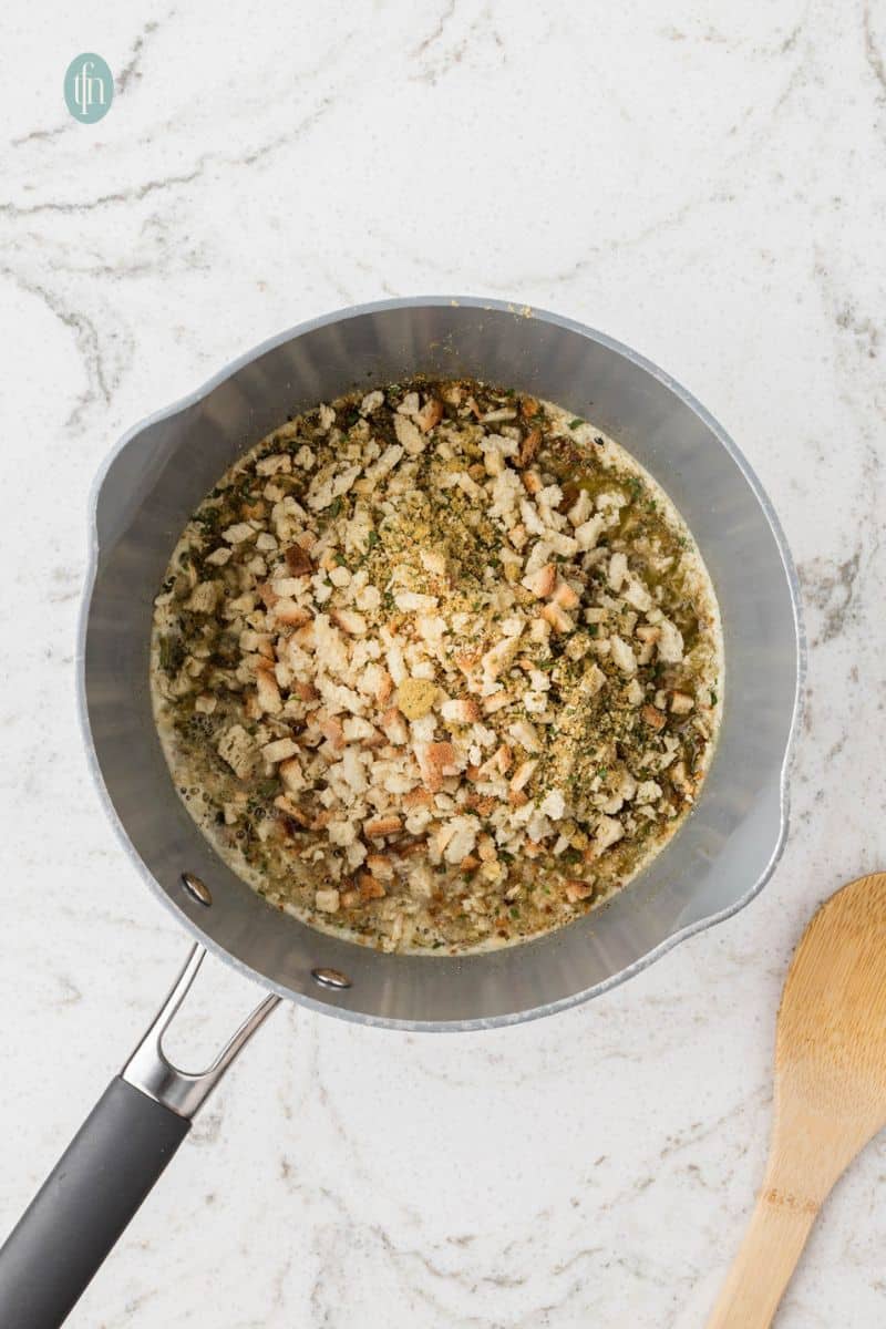 An overhead shot of a saucepan where small bread cubes and dried herbs have been added to a seasoned liquid, beginning to form the stuffing mixture.