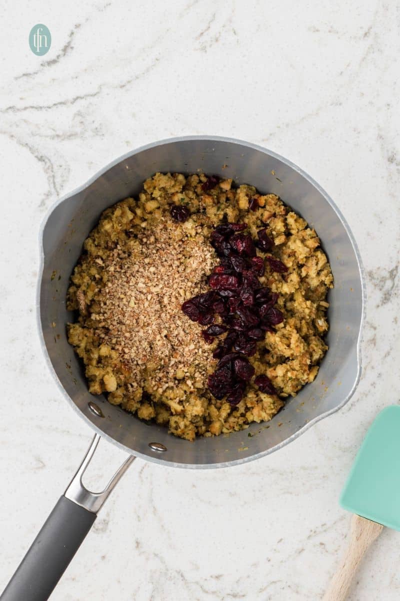 An overhead shot of a saucepan filled with the stuffing mixture, topped with dried cranberries and crushed pecans or walnuts, ready to be mixed.